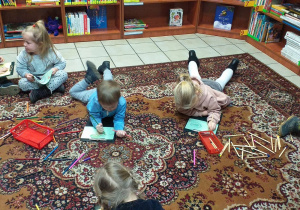 Dzieci leżąc na dywanie w bibliotece rysują na kartkach.