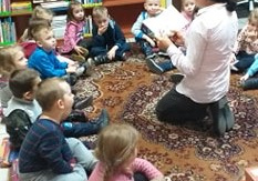 Dzieci siedza na dywanie i słuchają wierszy o babci i dziadku czytanych przez panią z biblioteki.